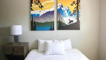 Habitación de hotel con cama, almohadas blancas, mesita de noche con lámpara y dos cuadros con paisajes de montañas y fauna en la pared.