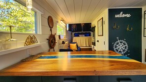 Um quarto com uma mesa de madeira, uma cama com cabeceira azul e uma bússola fixada na parede, além de uma placa de boas-vindas.