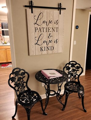 Una habitación con un tapiz de madera que reza 'Love is PATIENT & Love is KIND', una pequeña mesa redonda con un libro y dos sillas con diseños elaborados.