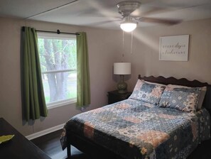 A bedroom with a bed, a nightstand, a lamp, a ceiling fan, and a window with curtains.