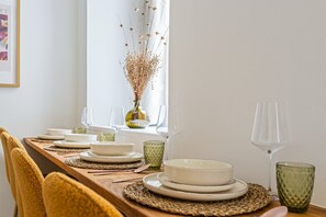 Ett matbord dukat med tallrikar, skålar och glas, med en vas med torkade blommor och ett fönster i bakgrunden.