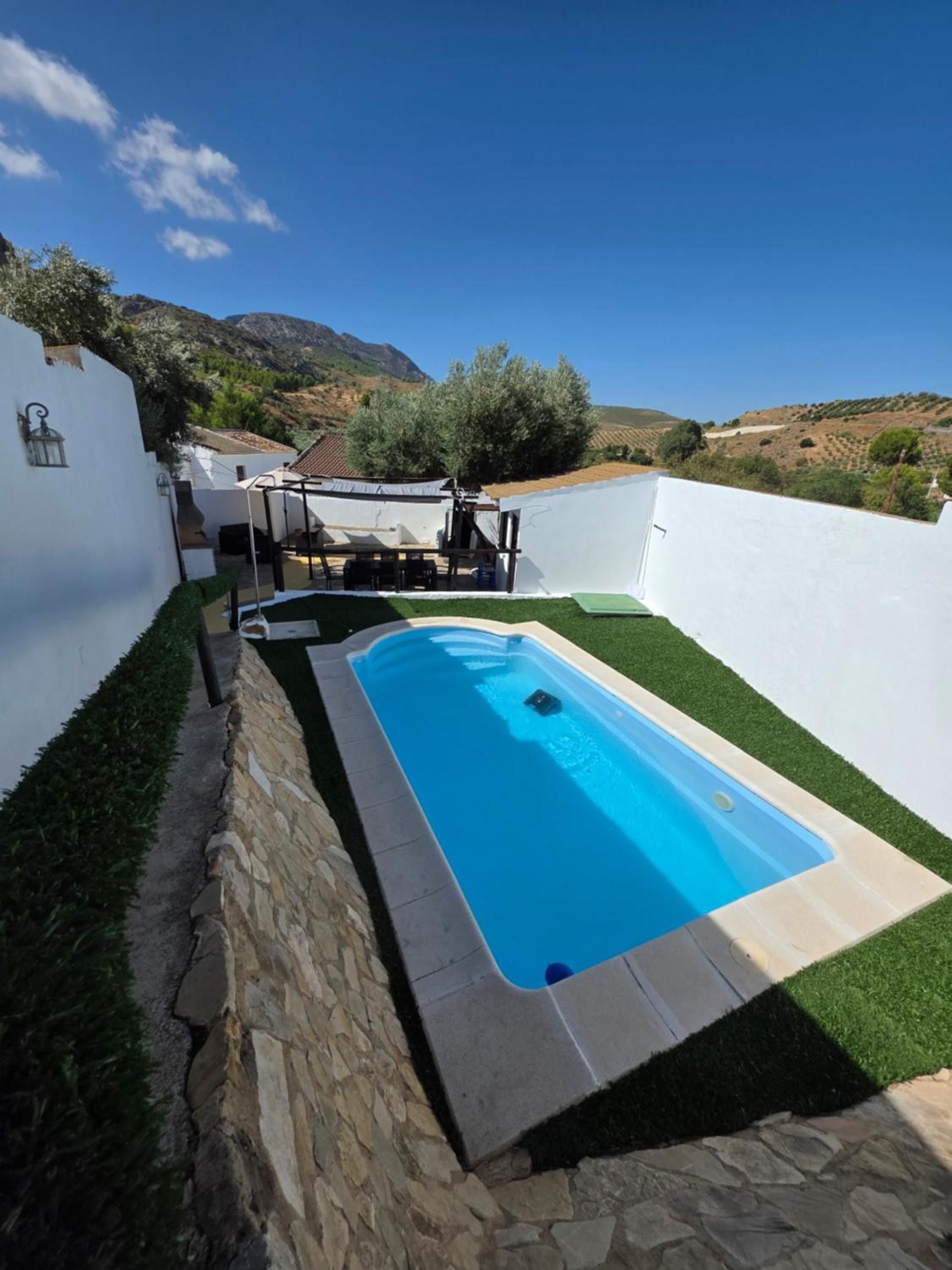 Une piscine bordée d’un mur en pierre et entourée de gazon synthétique, le tout adossé à un bâtiment blanc et offrant une vue sur des collines.
