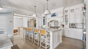 A modern kitchen with white cabinetry, a central island with bar stools, pendant lighting, and a view of the dining area. - Luxury Southold escape featuring a private pool, movie theater, gym, and... (Southold)