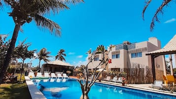 A swimming pool with a potted plant in the foreground, surrounded by palm trees and a building with a tiled roof in the background.