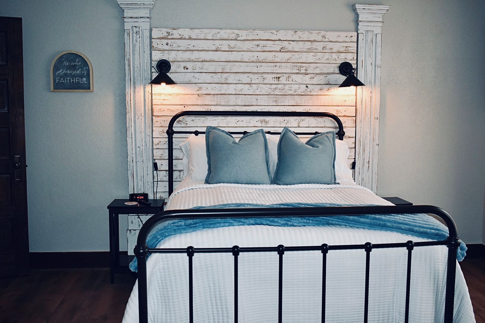 Ein Bett mit Metallgestell, weißer Bettwäsche und zwei Kissen. Über dem Bett hängt eine Wandlampe und daneben steht ein kleiner Tisch.