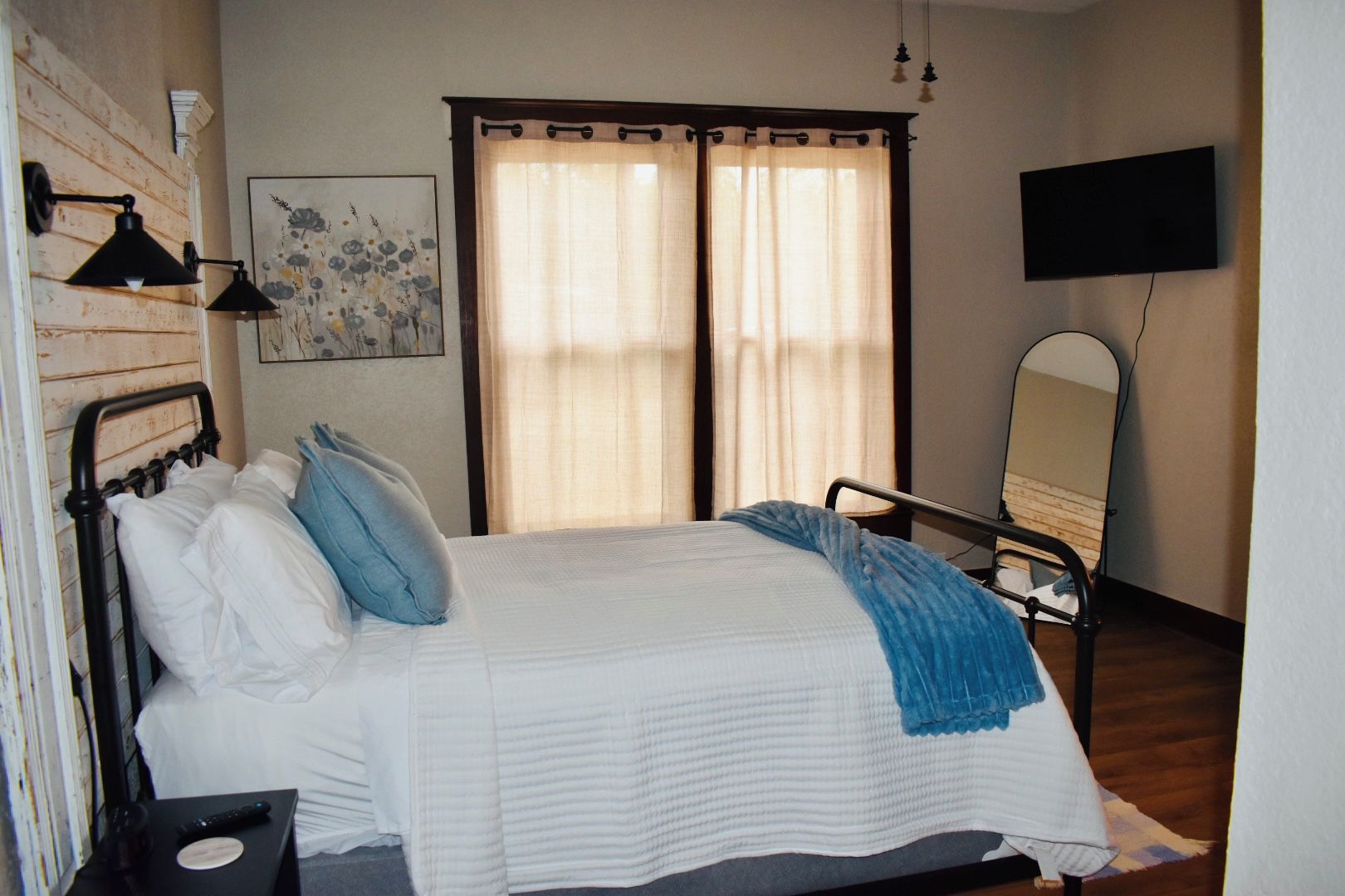A hotel room with a bed, bedside table, lamp, curtains, a painting, a television, and a mirror.