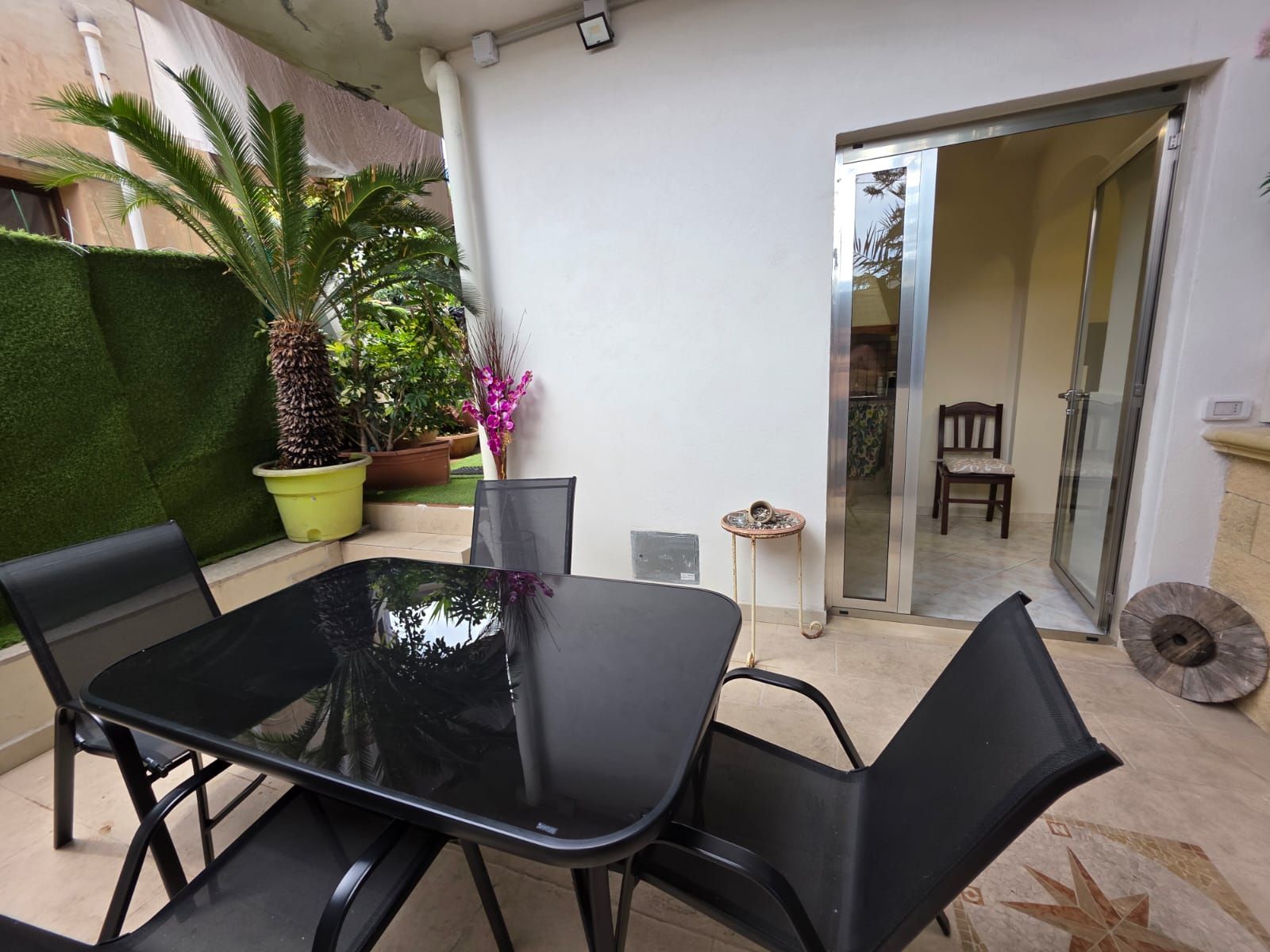Un patio con un tavolo e sedie in vetro, circondato da piante in vaso e un piccolo tavolino rotondo.