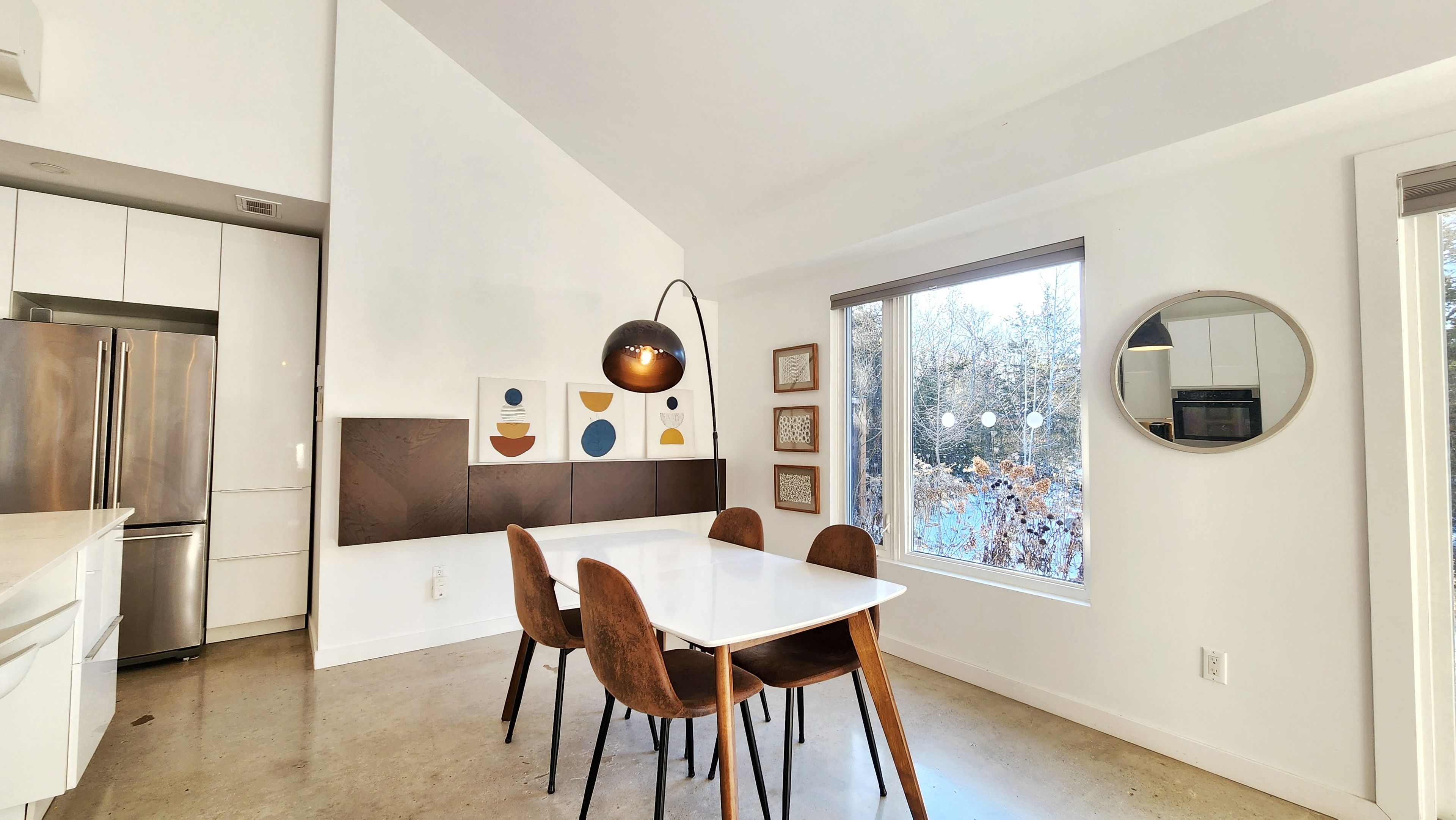 A modern kitchen with a built-in refrigerator, a dining area with a white table and brown chairs, and a large window with a view of trees.