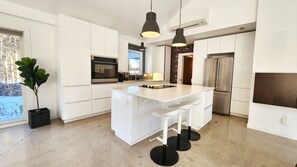 A modern kitchen with white cabinetry, a central island with bar stools, stainless steel appliances, pendant lighting, and a large window. - Luxury Cottage - Detox & Relaxation (Stone Mills)