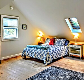 Une chambre avec un plafond en pente, un lit avec une literie à motifs, deux lampes de chevet, une fenêtre avec des stores et un petit cadre photo accroché au mur.