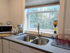 Une cuisine équipée d’un évier à deux bacs, d’un four à micro-ondes, d’une machine à café et d’une fenêtre donnant sur la verdure.