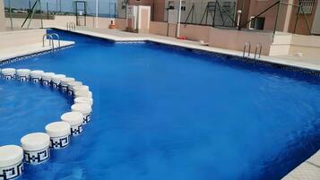 Ein Schwimmbecken mit weißen, kreisförmigen Stufen, die zum Wasser führen, umgeben von einem Zaun und einem Gebäude mit vielen Fenstern.