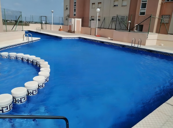 Une piscine avec une série de marches blanches et circulaires menant à l’eau, entourée d’une clôture et bordée d’un bâtiment doté de nombreuses fenêtres.