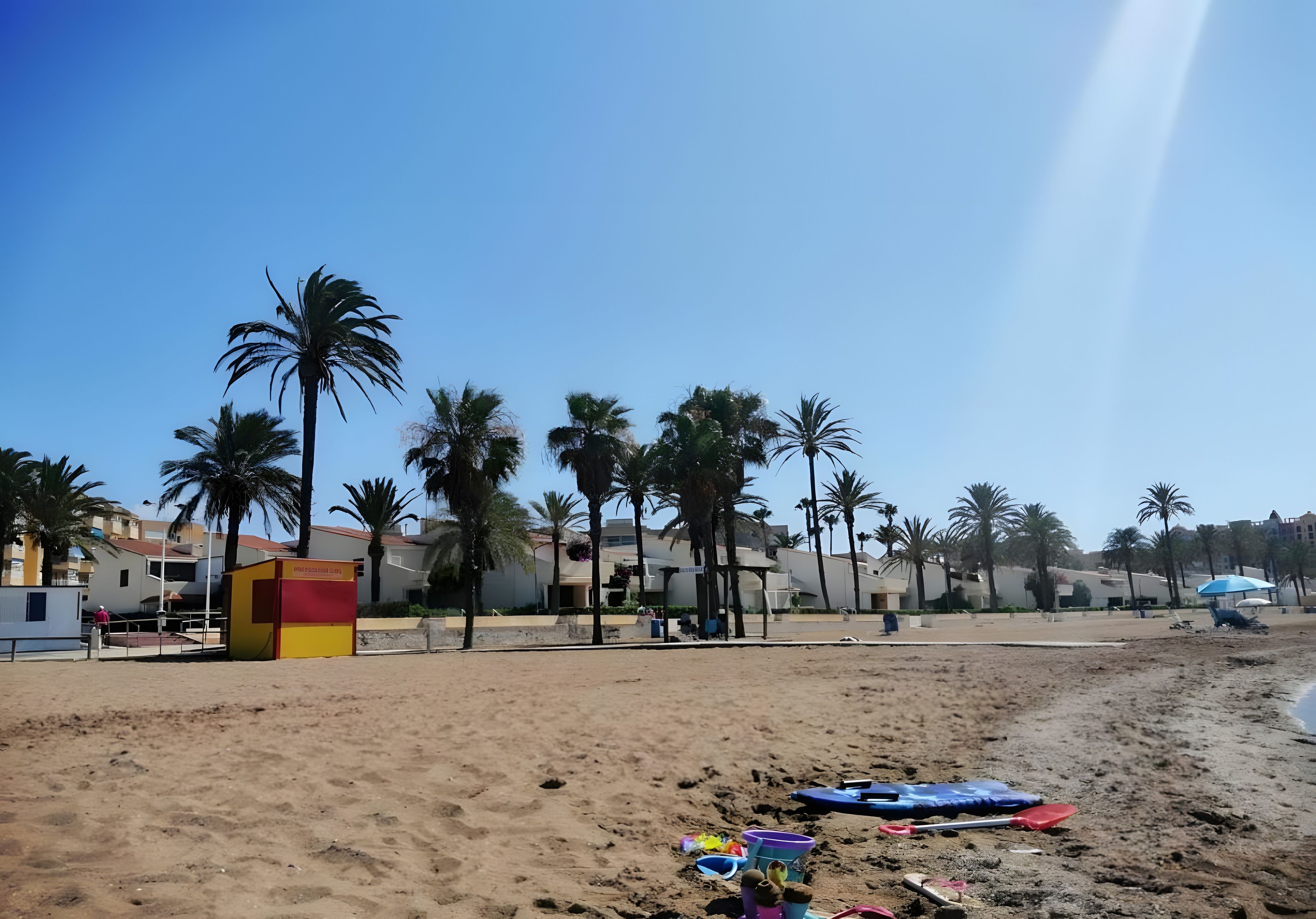 Une plage de sable bordée de palmiers, avec une tour de surveillance pour les sauveteurs et des bâtiments en arrière-plan.