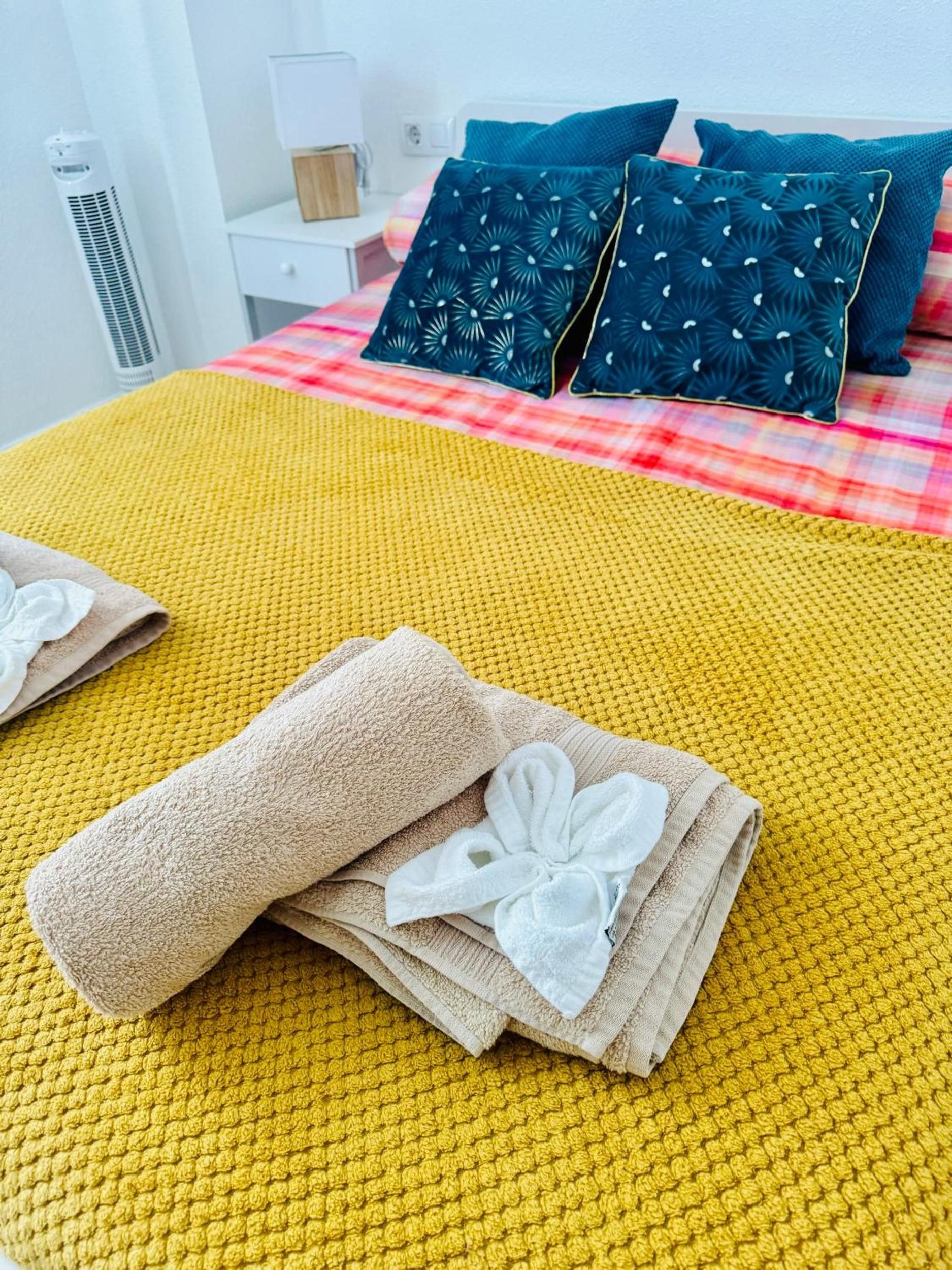 Un lit recouvert d’une couvre-lit jaune à motifs, deux oreillers ornés d’un motif de feuilles et deux serviettes pliées décorées d’une fleur blanche.