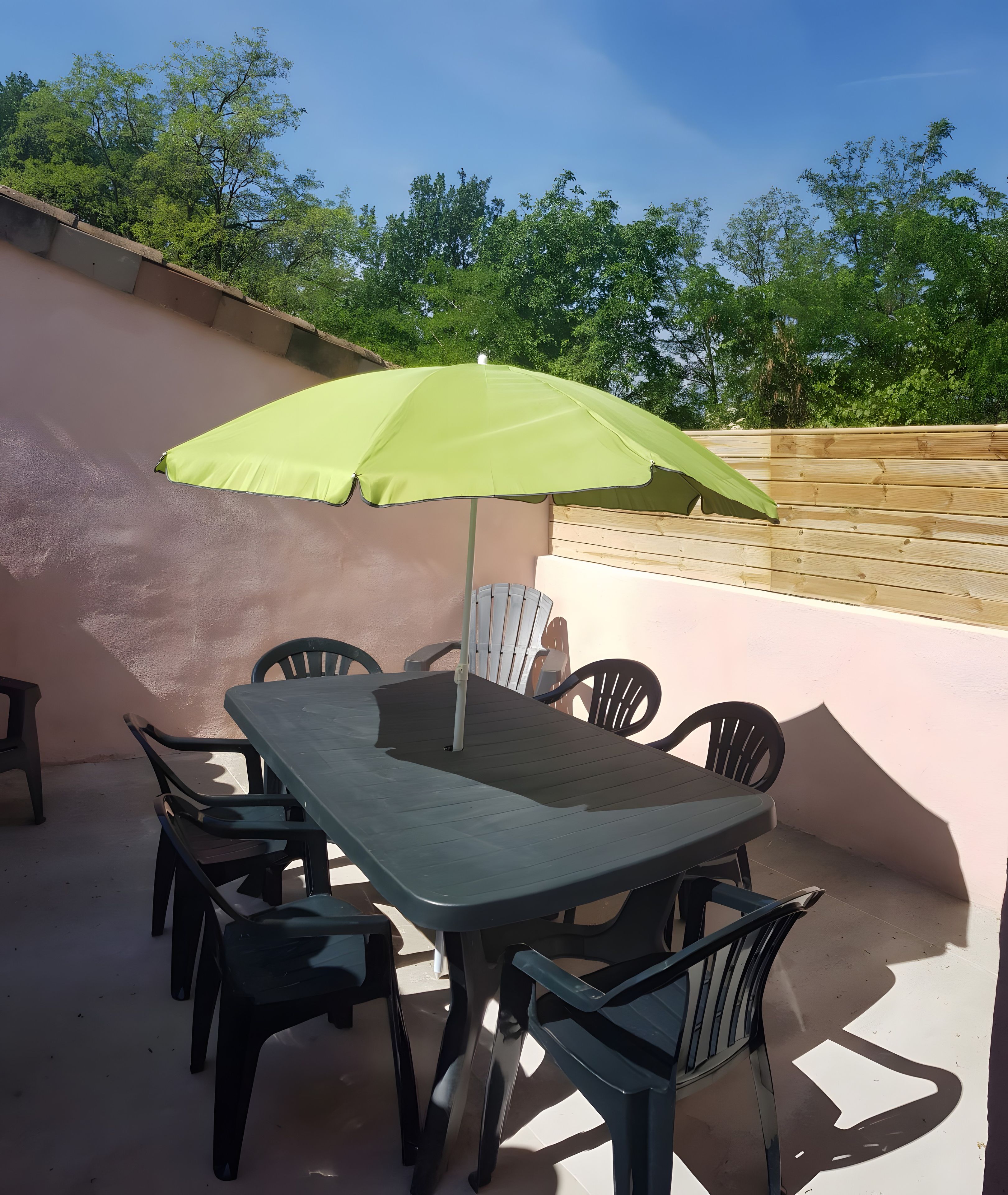 Een terras buiten met een tafel en stoelen, een groene parasol, en uitzicht op bomen en lucht.