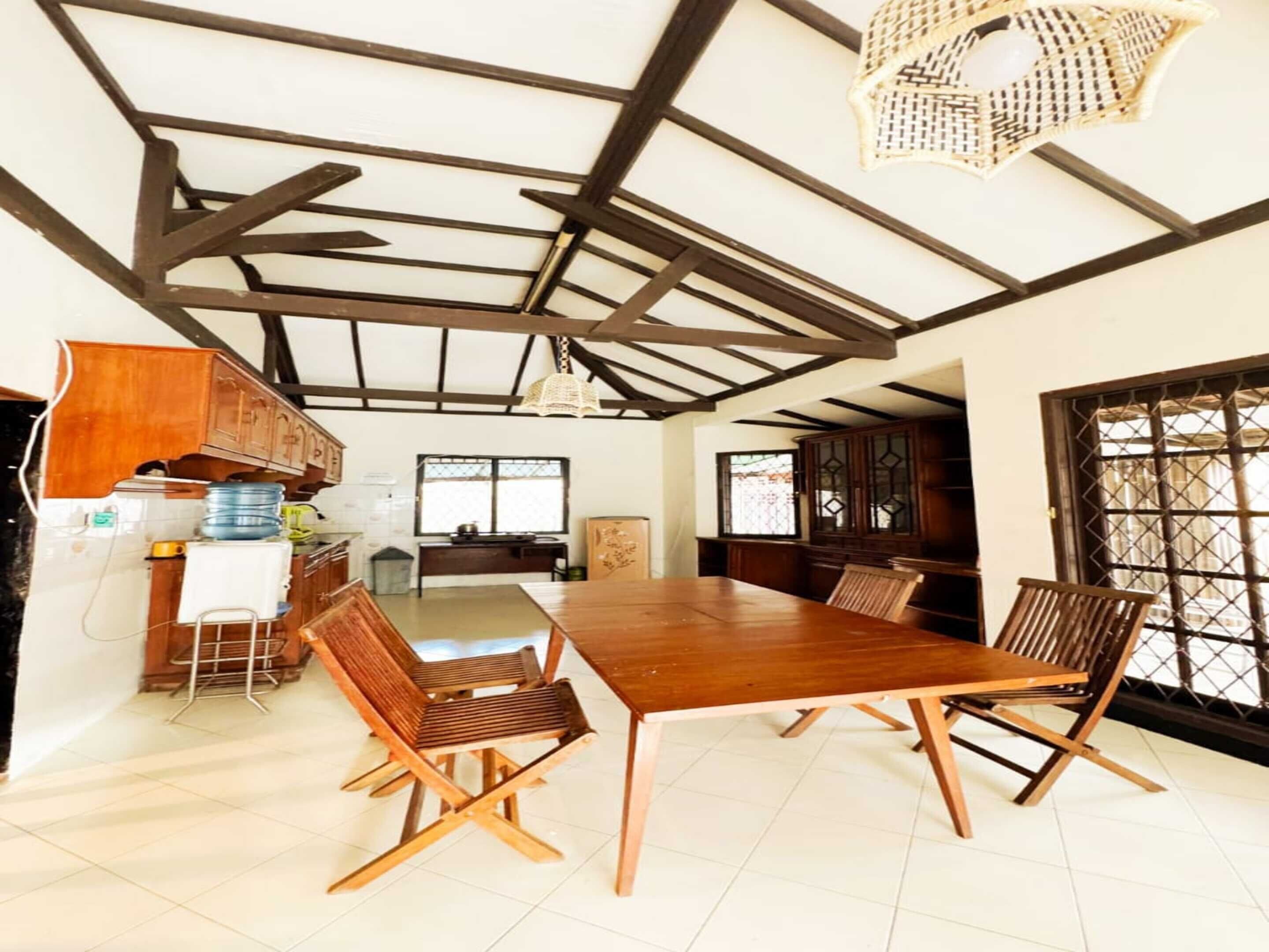A spacious dining area with a long wooden table and chairs, a kitchen area with cabinets and a water dispenser, and a view through a window with a lattice design.