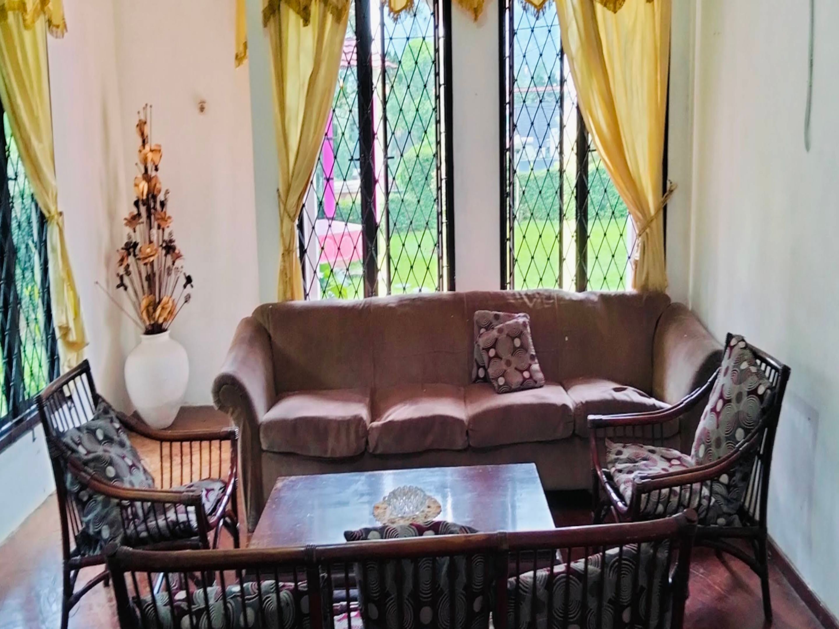 A living room with a brown sofa, two patterned armchairs, a glass coffee table, and a vase with dried flowers.