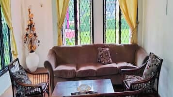 A living room with a brown sofa, two patterned armchairs, a glass coffee table, and a vase with dried flowers.