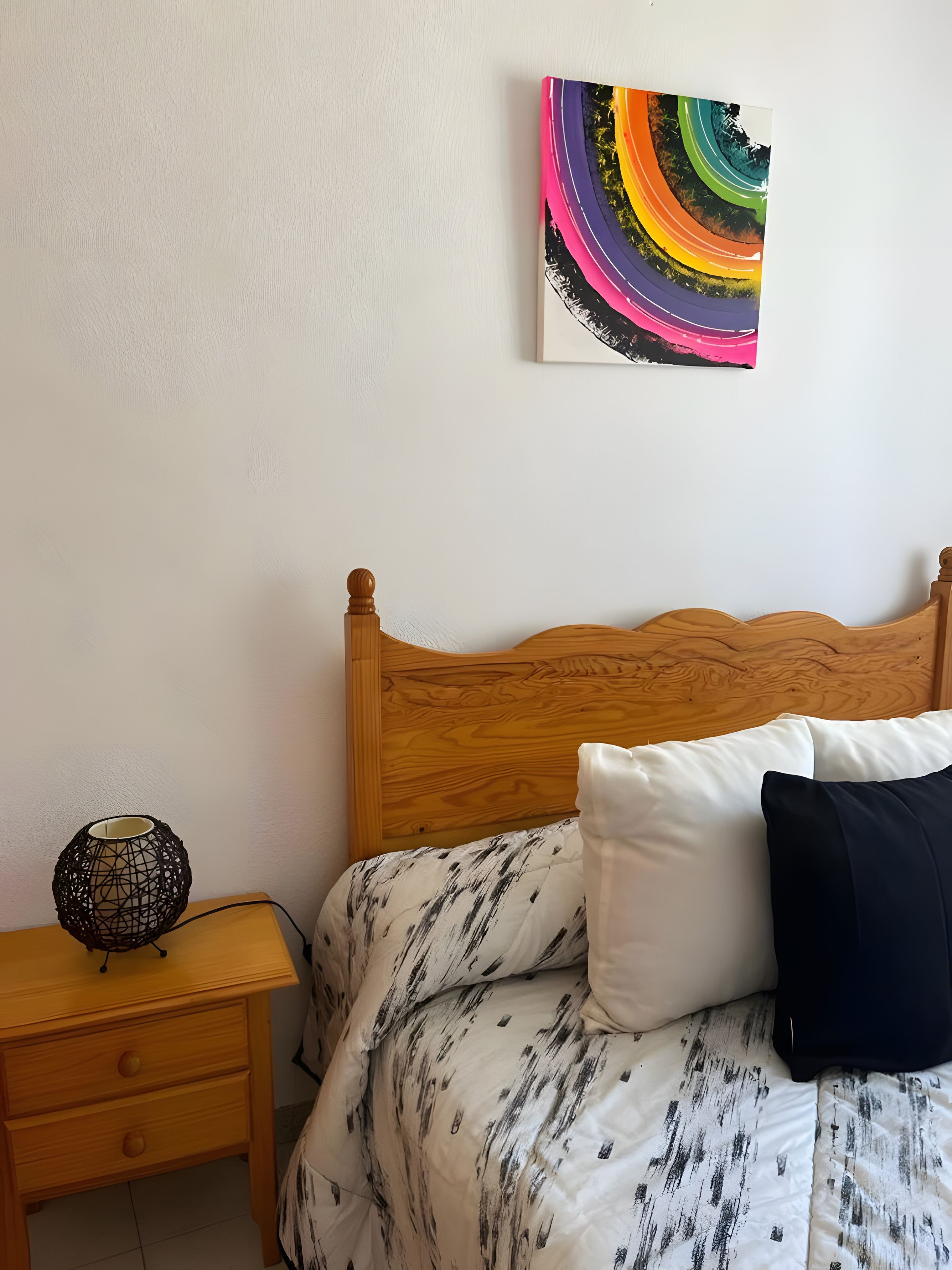 A bedroom with a wooden headboard, a bed with patterned bedding, a bedside table with a decorative lamp, and a colorful abstract painting on the wall.