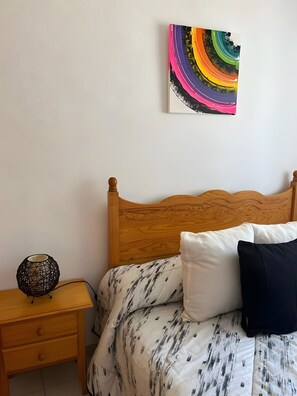 Ein Schlafzimmer mit einem hölzernen Kopfteil, einem Bett mit gemusterten Textilien, einem Nachttisch mit einer dekorativen Lampe und einem farbenfrohen abstrakten Gemälde an der Wand.
