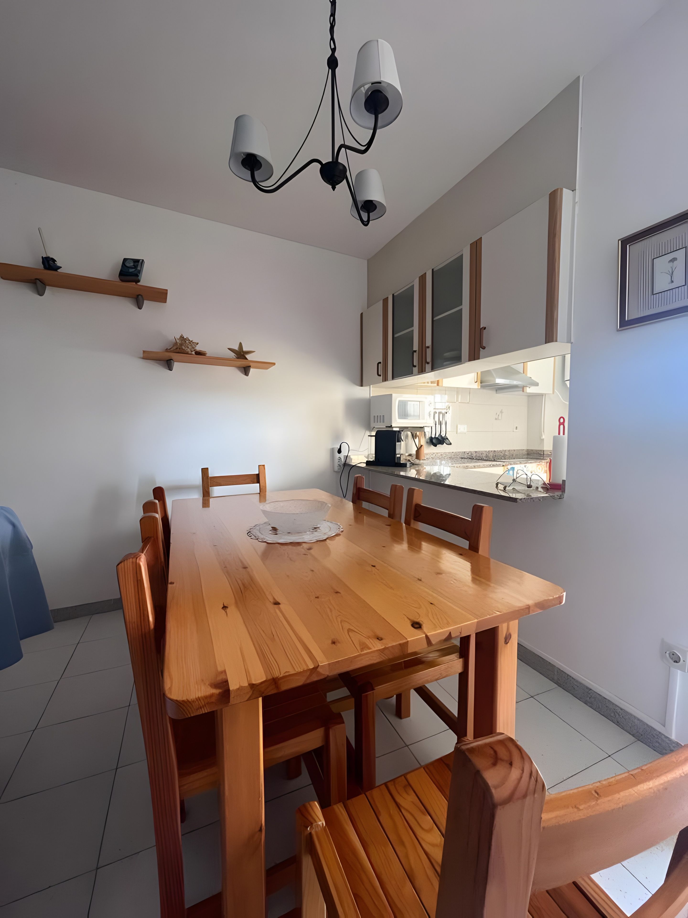 A dining area with a wooden table and chairs, a chandelier, and a kitchen in the background.