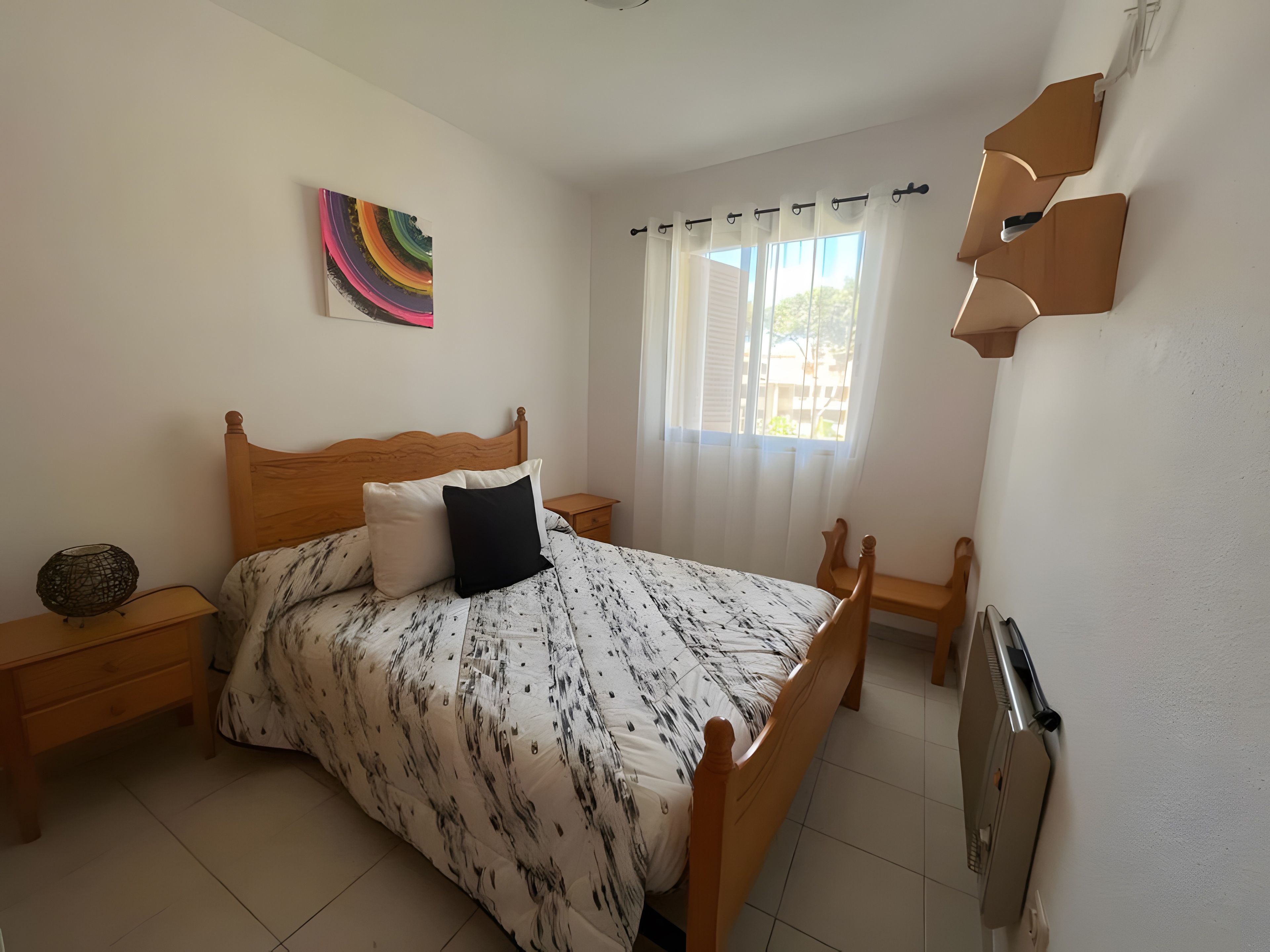 Un dormitorio con cama de madera, mesita de noche, lámpara, ventana con cortinas y radiador fijado a la pared.