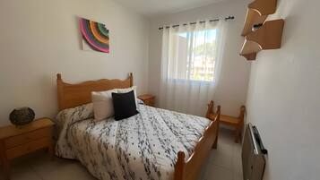Un dormitorio con cama de madera, mesita de noche, lámpara, ventana con cortinas y radiador fijado a la pared.