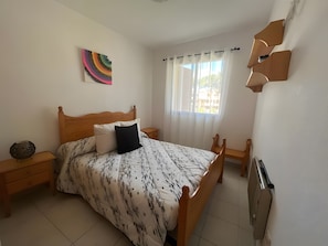 A bedroom with a wooden bed, a nightstand, a lamp, a window with curtains, and a wall-mounted radiator.