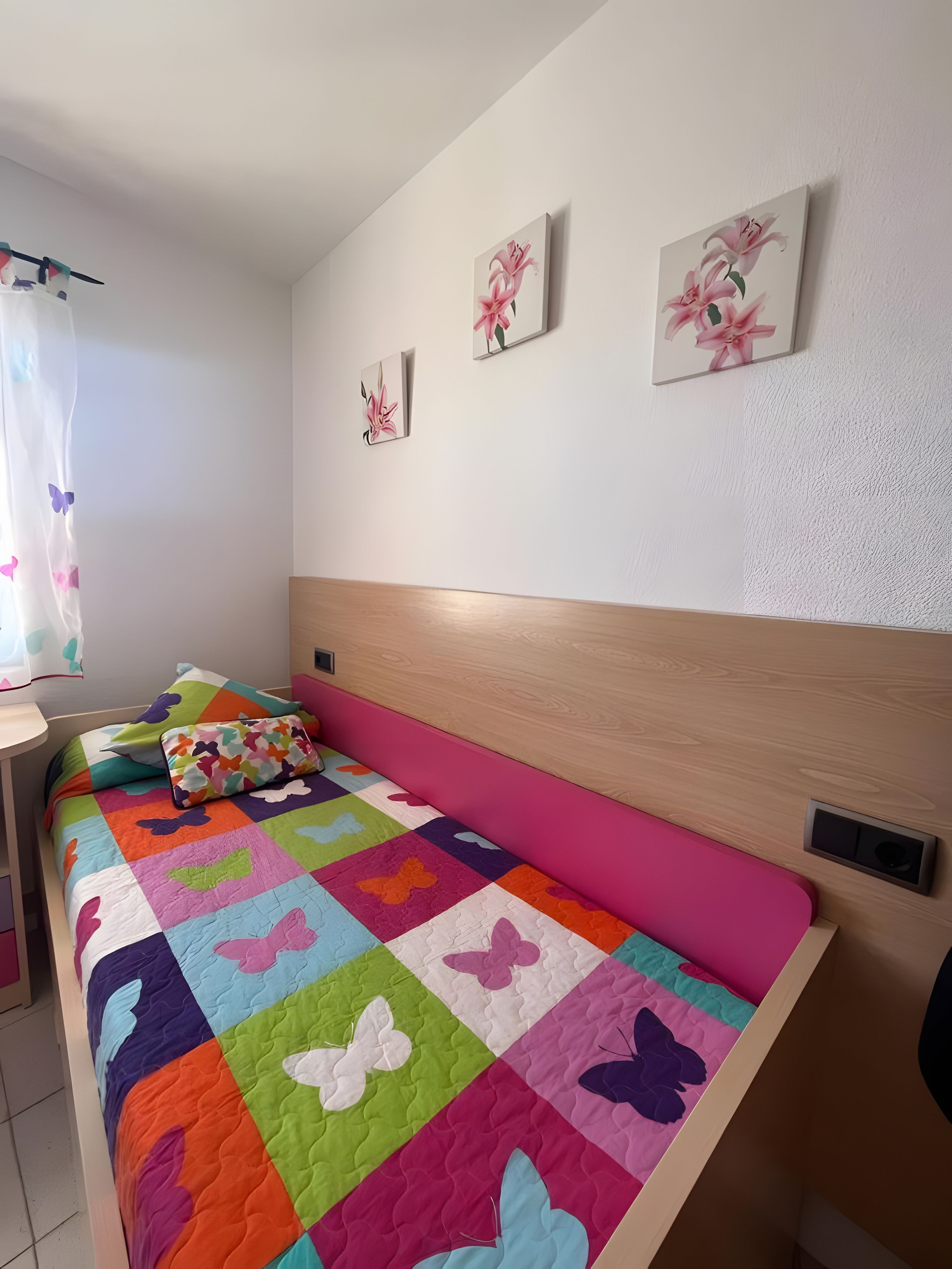Quarto com colcha colorida, cabeceira de madeira e três quadros florais fixados na parede.