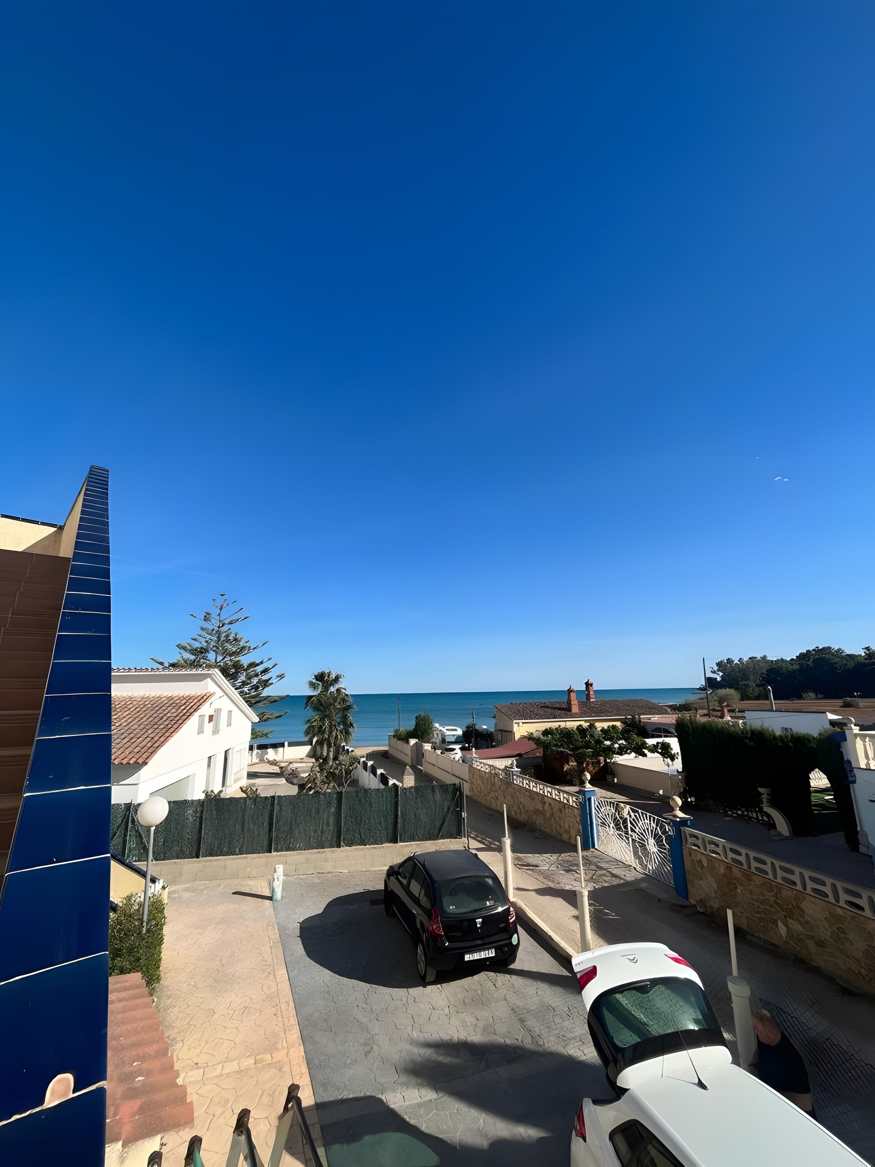 Área residencial com vista ampla para o mar, um carro preto na entrada e um carro branco em primeiro plano.
