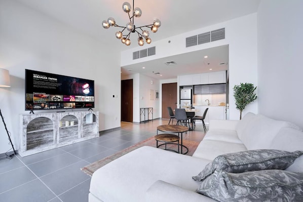 A modern living room with a flat-screen TV, a white sofa, a dining area, and a kitchen in the background. - Chic 1BR Escape in Creek Edge | Creek Harbour (Dubai)