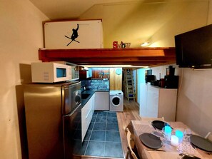 A compact kitchen with a refrigerator, microwave, and washing machine. A dining area with a table set for two is visible. - Arc 2000 Splendide 5* (Renovated) Ski-In/Ski-Out, Wi-Fi (Bourg-Saint-Maurice)