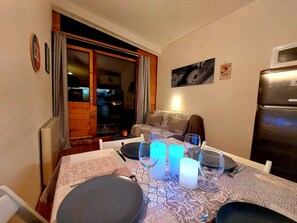 Ein Essbereich mit Tisch für zwei Personen, einer Couch, einem Kühlschrank und einer Glasschiebetür, die auf einen Balkon führt.