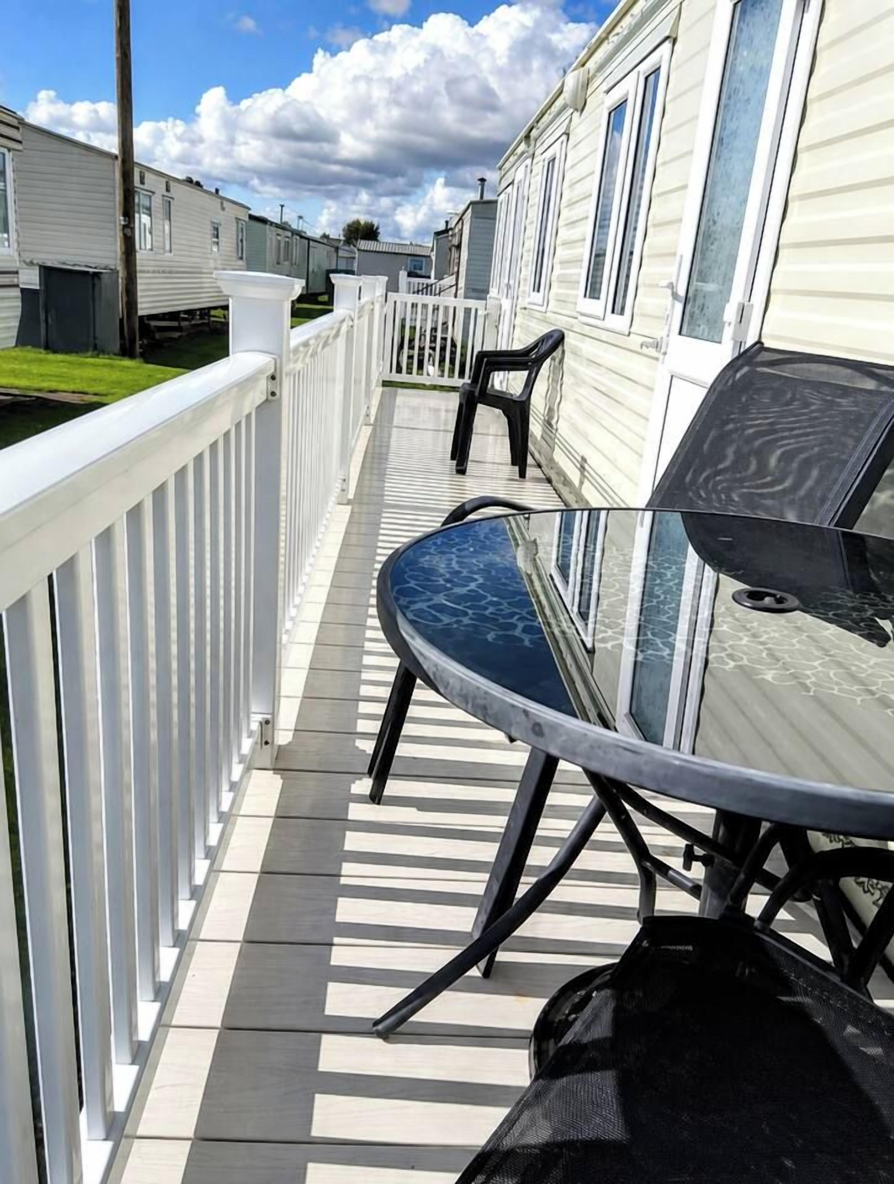 A deck with a table and chairs, surrounded by a white railing, with mobile homes in the background.