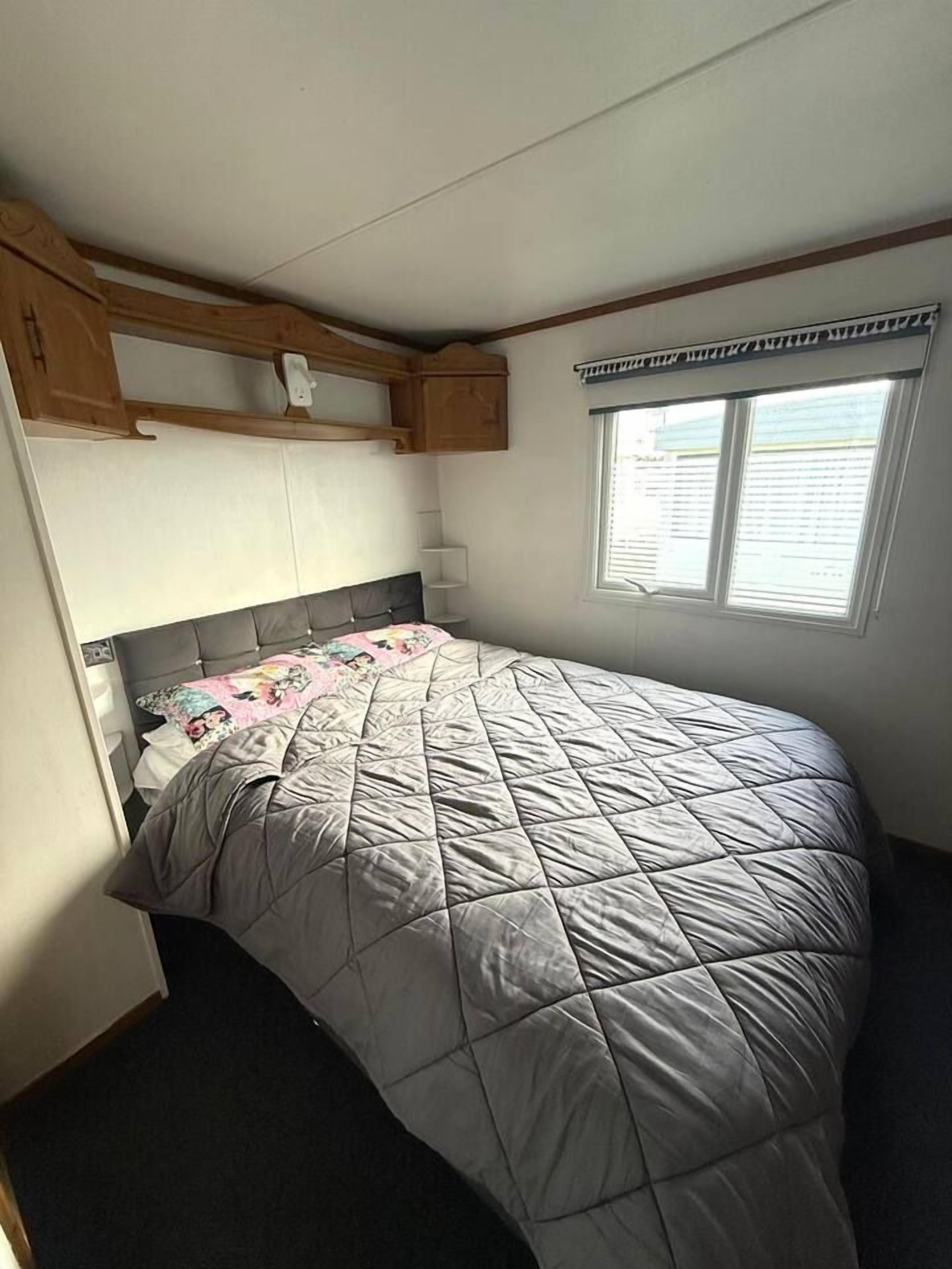 A single bed with a quilted grey bedspread, a headboard, and a window with blinds.