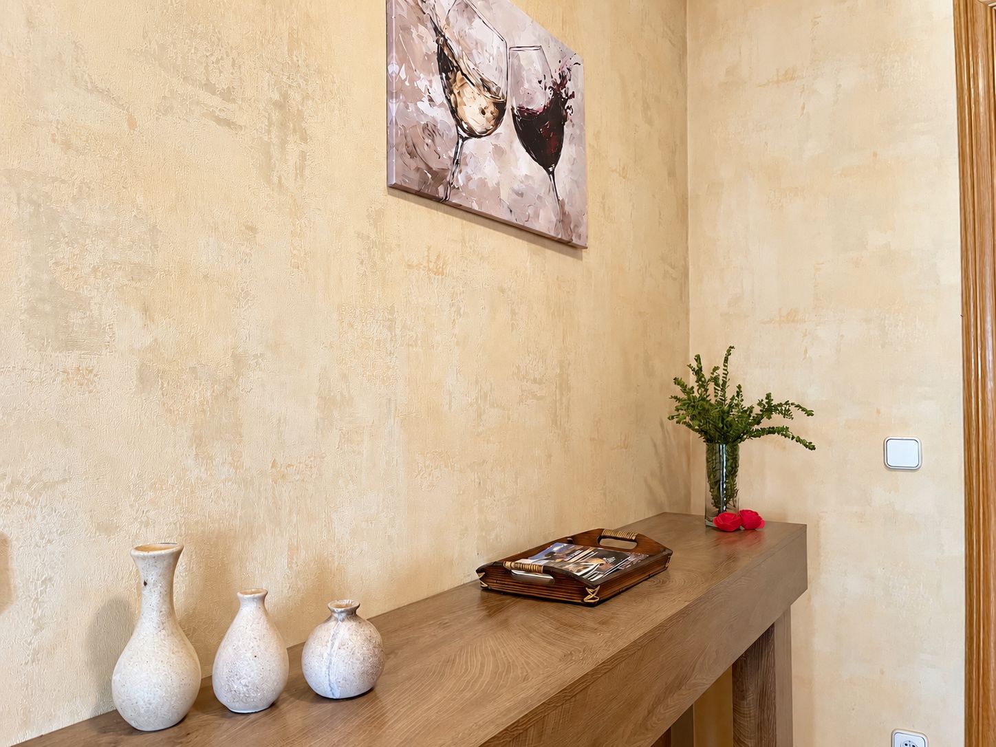 A reception area with a wooden counter, three white vases, a magazine rack, a painting of wine glasses, a small plant, and a red flower.