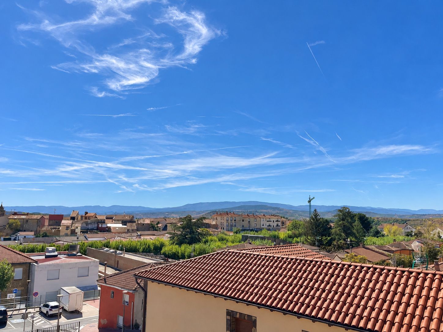 A residential area with terracotta-tiled roofs, a clear blue sky, and distant mountains.