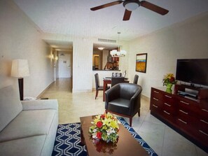 A living room with a sofa, a coffee table with flowers, a TV, and a dining area in the background. - DIVI VILLAGE GOLF & BEACH RESORT - 1 BEDROOM SLEEPS 4. STEPS AWAY FROM THE BEACH (Oranjestad)
