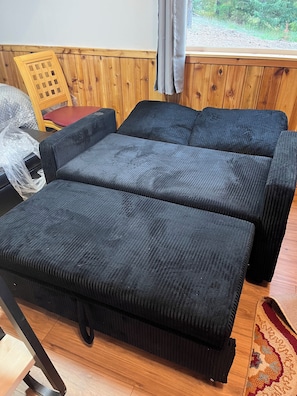 Ein Zimmer mit zwei schwarzen Polstersofas, einem Holzstuhl, Holzboden und einem Vorhangfenster.