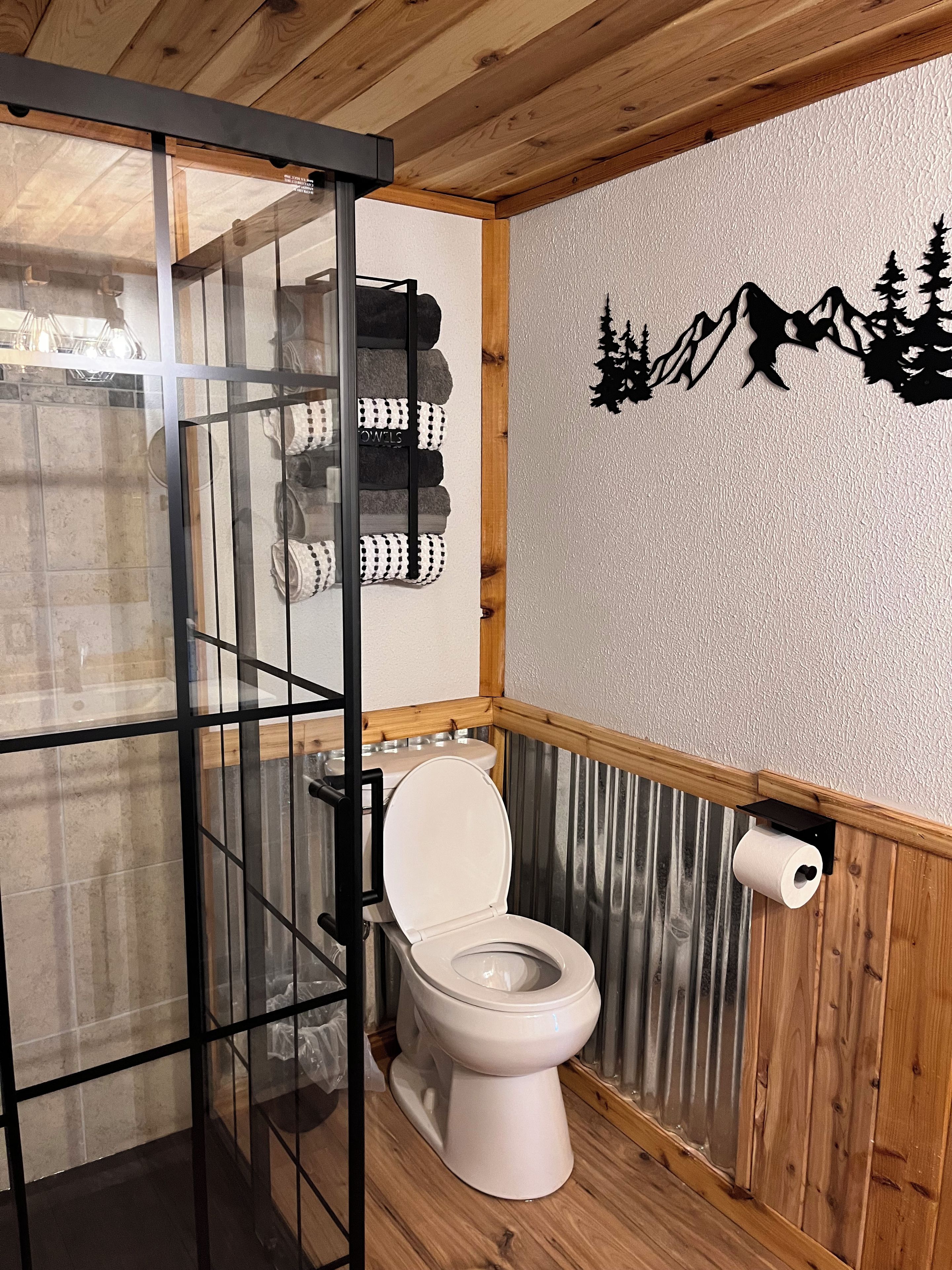 Une salle de bain avec un plancher en bois, des toilettes, une douche en verre et un motif mural représentant des montagnes et des arbres.