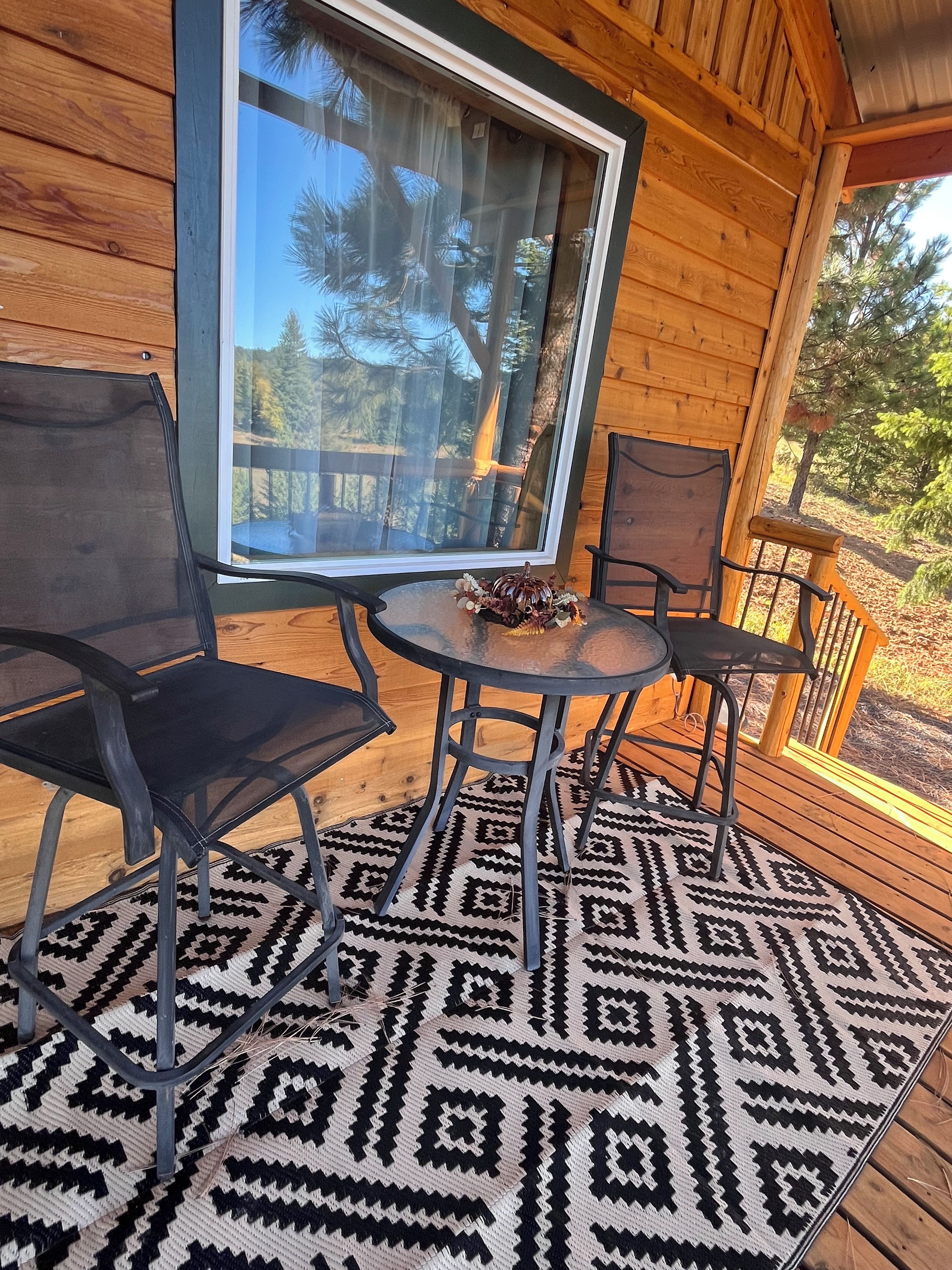 Terraza de madera con puerta de vidrio, dos sillas, una mesa y un tapete estampado.