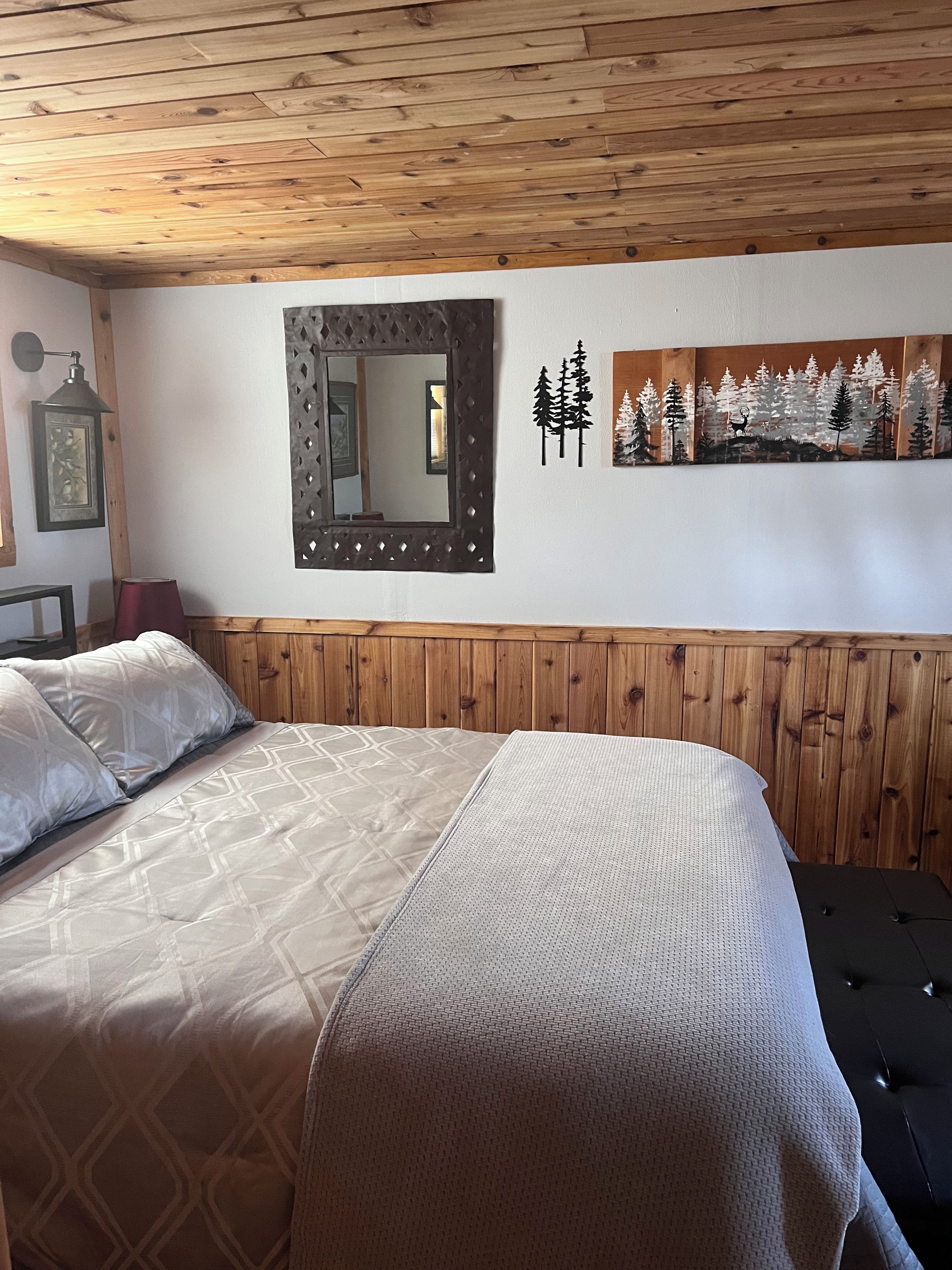 A bedroom with a wooden headboard, a bed with patterned bedding, a wall-mounted mirror, and a forest-themed artwork.