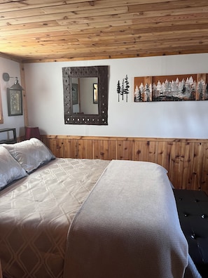 A bedroom with a wooden headboard, a bed with patterned bedding, a wall-mounted mirror, and a forest-themed artwork.