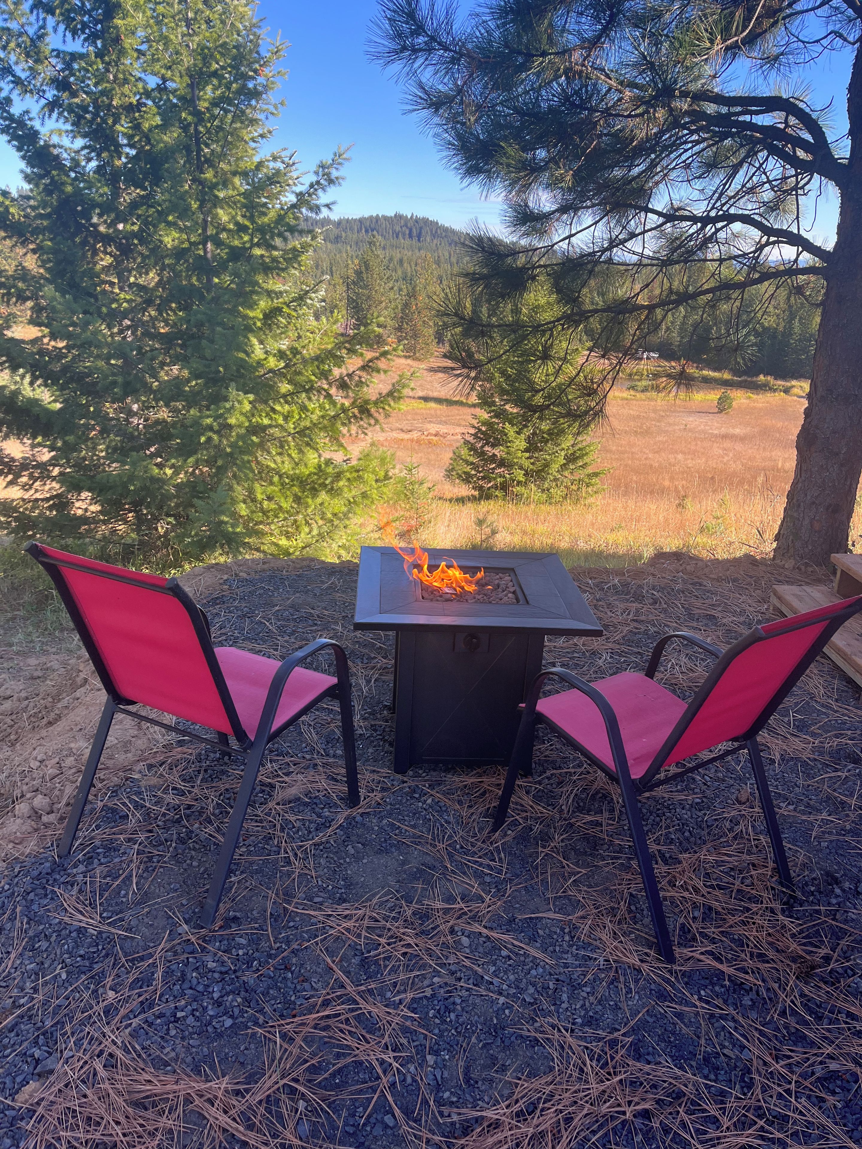A fire pit table with two red chairs, surrounded by pine trees and a view of a forested area.