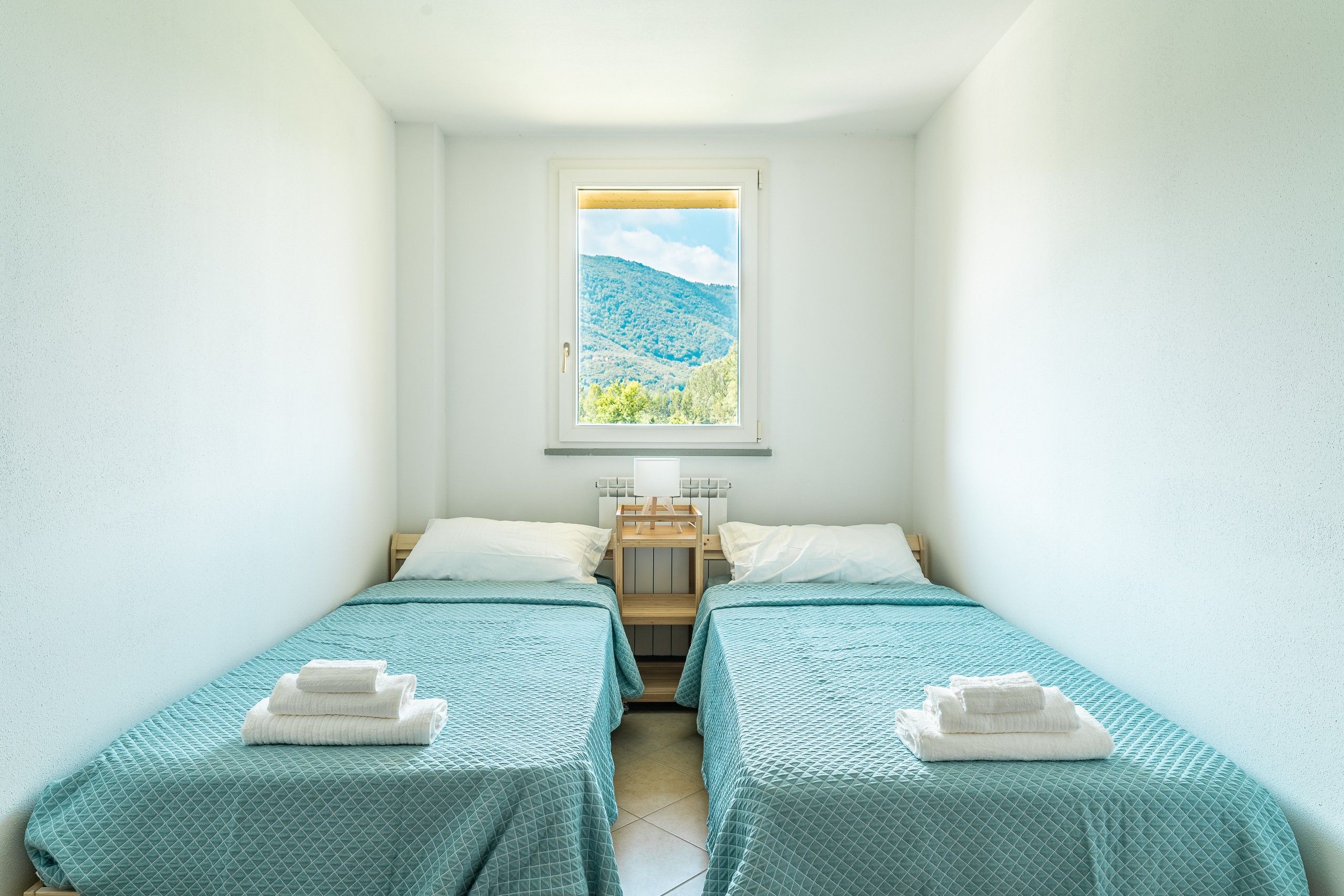 A small, clean room with two beds, a window with a mountain view, and a bedside table with a lamp.