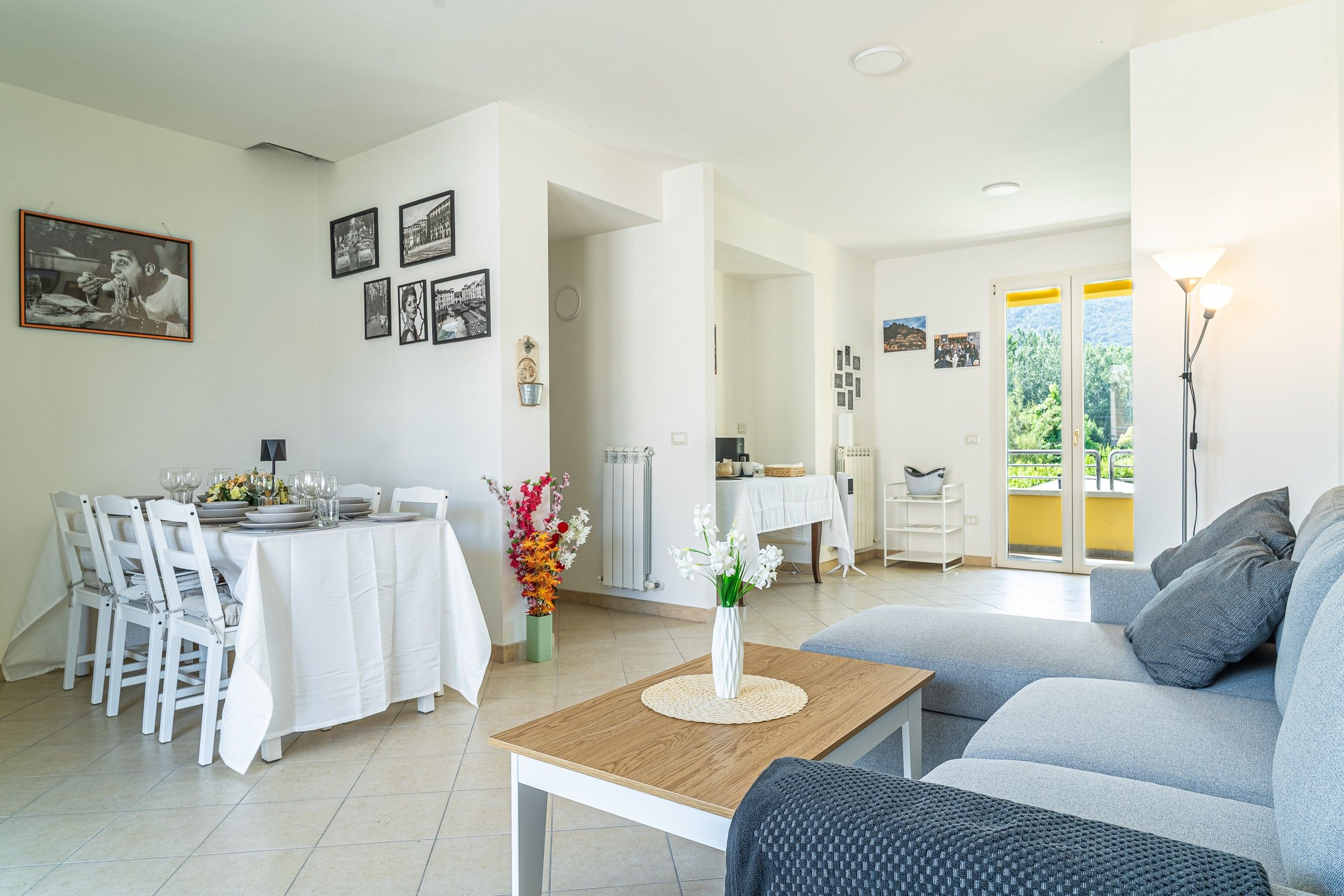 Ein modernes Wohnzimmer mit Essbereich, ausgestattet mit einer Couch, einem Esstisch, Stühlen und einem Couchtisch. An den Wänden befinden sich gerahmte Bilder und durch eine Glastür ist ein Blick ins Freie möglich.