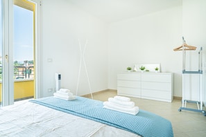 A hotel room with a bed, a white dresser, a drying rack, and a view of the outdoors.