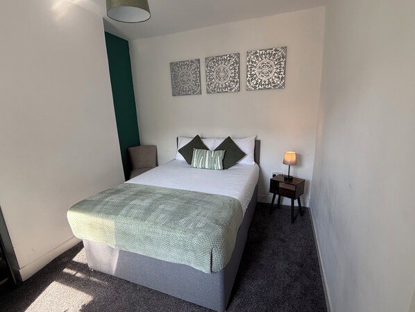 Una cama bien hecha con una manta verde, una lámpara de noche y decoración en la pared.