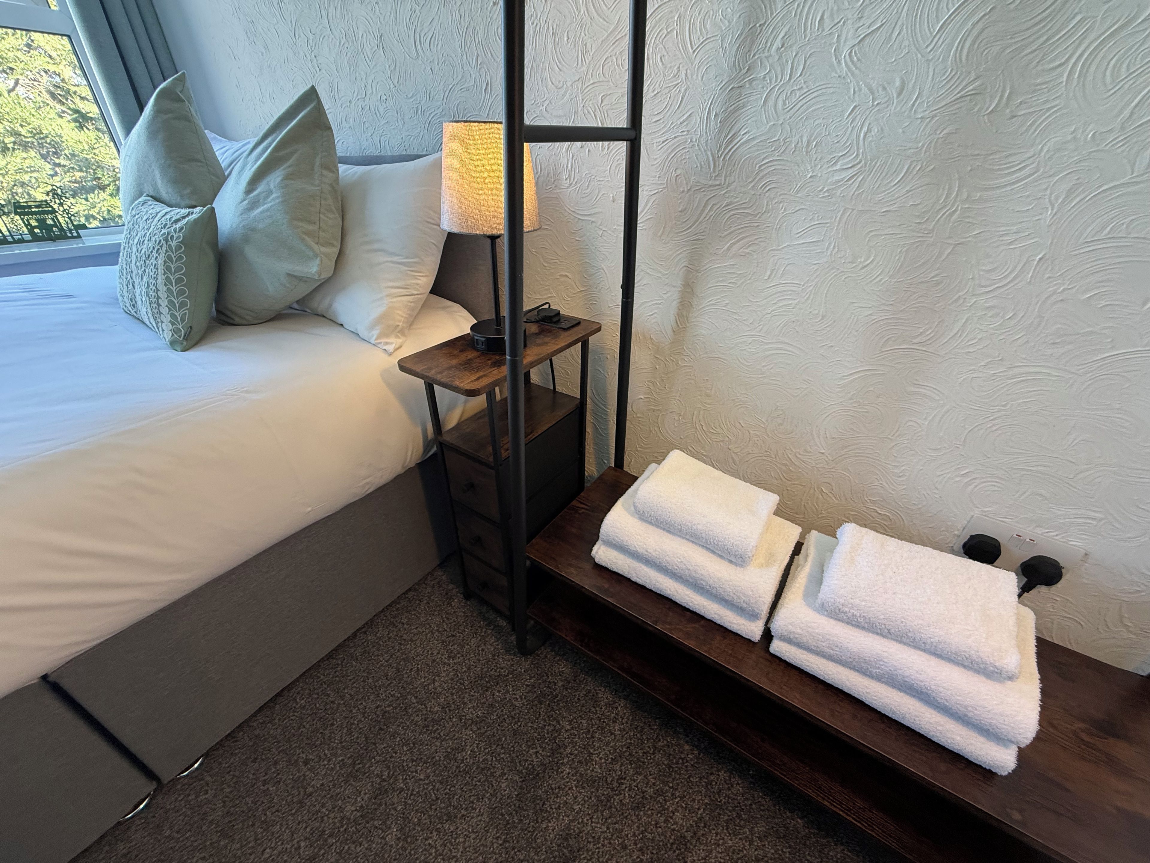 A neatly made bed with pillows, a bedside lamp, and a shelf with folded towels.